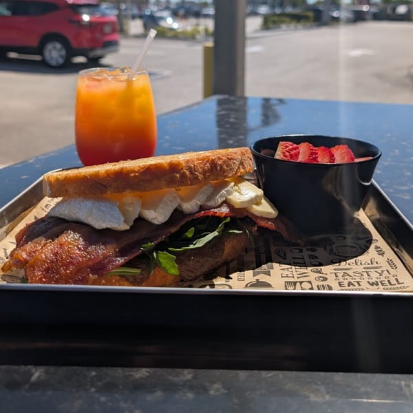 A breakfast sandwich with bacon, eggs, and arugula, served with fruit and an orange drink.