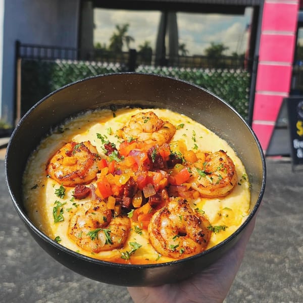 A hand holds a black bowl of shrimp and grits, topped with diced vegetables and parsley.