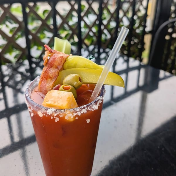 A Bloody Mary cocktail garnished with bacon, olives, cheese, and pickles on a patio table.