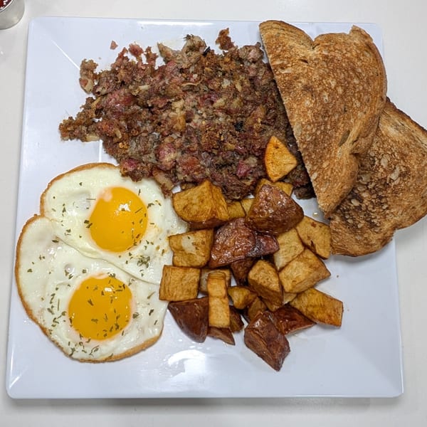 A breakfast plate with two fried eggs, corned beef hash, roasted potatoes, and two slices of toast.