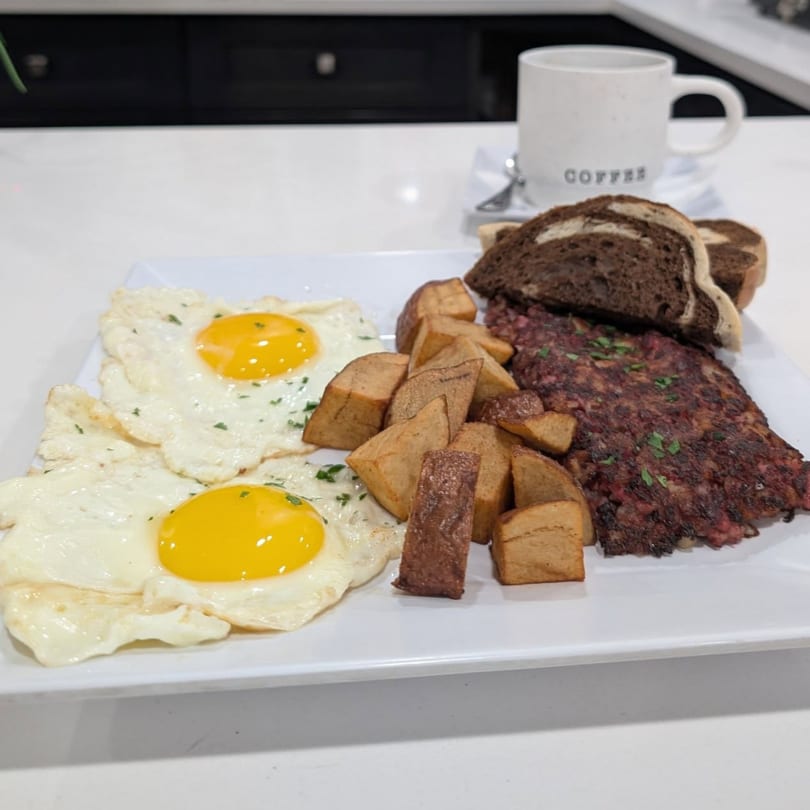 Breakfast plate with two fried eggs, home fries, corned beef hash, and marble rye toast.